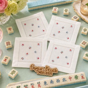May include: Four white square fabric napkins featuring embroidered Mahjong tile motifs in red, green, and blue. Each napkin has a stitched border. A wooden sign with the text Sugar Beach EMBROIDERY is placed in the center among Mahjong tiles.