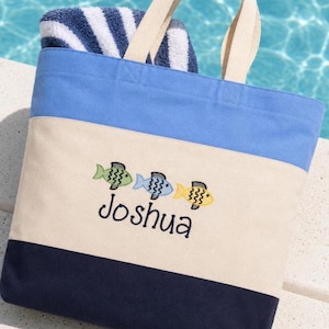 May include: A color-blocked tote bag featuring horizontal stripes of light blue, cream, and navy blue. The cream section displays an embroidered design of three small fish in green, blue, and yellow, with the name Joshua stitched in dark blue.