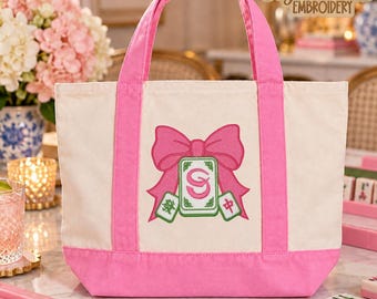 May include: A cream-colored canvas tote bag with bright pink handles and a pink base. The front features an embroidered design of a large pink bow centered above three mahjong tiles, with the center tile displaying the letters SG.