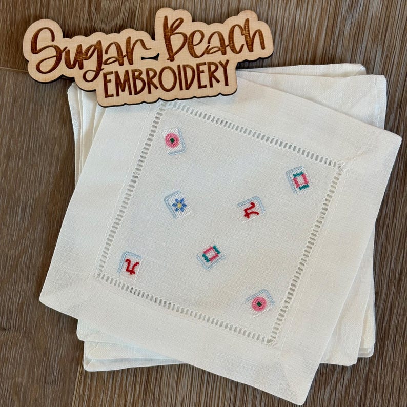 May include: A stack of white square linen napkins featuring a hemstitched border and small embroidered mahjong tile motifs in various colors. A wooden sign with the text Sugar Beach EMBROIDERY is positioned above the napkins.