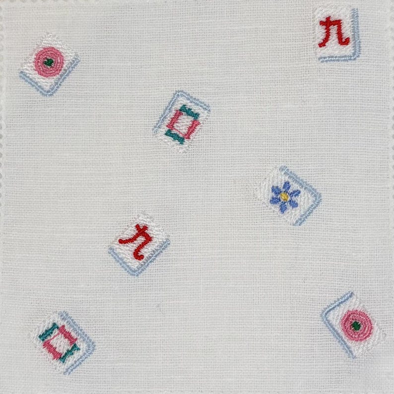 May include: A white fabric square featuring scattered embroidered mahjong tile motifs. The designs include red Chinese characters, pink circles, blue flowers, and green and pink rectangular patterns, all outlined in light blue stitching.