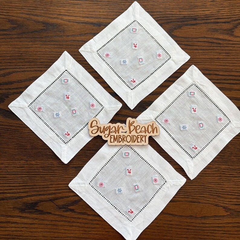 May include: Four square white linen cocktail napkins featuring a hemstitched border and scattered small embroidered motifs in red, blue, and green. A wooden sign in the center displays the text Sugar Beach Embroidery in script.
