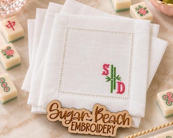 May include: A stack of white linen cocktail napkins featuring a hemstitched border and a custom monogram with the letters S and D in pink, separated by a green bamboo stalk. A wooden sign reads Sugar Beach Embroidery.