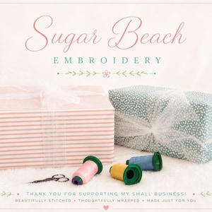 May include: Two rectangular gift boxes wrapped in white tulle ribbon, one with pink stripes and one with a green floral pattern. Three spools of thread in pink, yellow, and blue sit beside a pair of small scissors. Text reads Sugar Beach Embroidery.