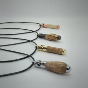 Può includere: Quattro collane con pendente caratterizzate da corpi cilindrici in legno con tappi e punte metalliche. Ogni pendente è fissato a un sottile cordino nero. I componenti metallici variano nel colore, includendo finiture oro rosa, canna di fucile, oro e argento.