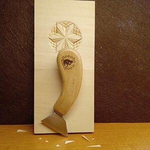 May include: A wood carving tool with a curved wooden handle and a metal blade. The handle is stamped with the text "Bonobo Wood" and a logo. The tool is positioned on a light-colored wooden surface with a carved star design.