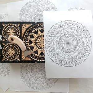 Puede incluir: Un bloque de tallado en madera negra y natural con intrincados patrones geométricos de estrellas, acompañado de una herramienta de metal con mango de madera clara etiquetada como Bonobo Wood y una hoja de papel blanco con un dibujo lineal circular.