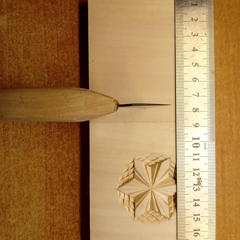 May include: A close-up of a wood carving tool and a carved wooden starburst design. A ruler with millimeter markings is next to the carving. The tool has a wooden handle and a sharp metal blade.