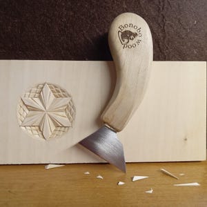 Puede incluir: Una herramienta para tallar madera con un mango de madera curvado y una cuchilla plateada, junto a una estrella de madera tallada. El mango está estampado con el texto "Bonobo Wood". Hay virutas de madera esparcidas por la superficie.