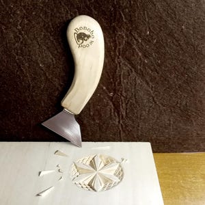 May include: A light-colored wood carving knife with a curved handle and a silver blade is shown in use. The handle has the text "Bonobo Wood" and a graphic. The knife is carving a star design into a light-colored wooden surface.