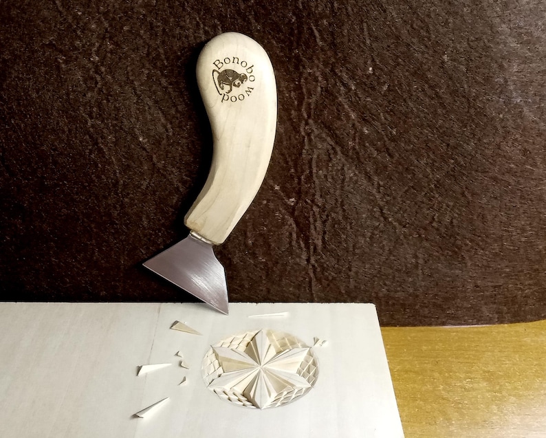 May include: A light-colored wood carving knife with a curved handle and a silver blade is shown in use. The handle has the text "Bonobo Wood" and a graphic. The knife is carving a star design into a light-colored wooden surface.