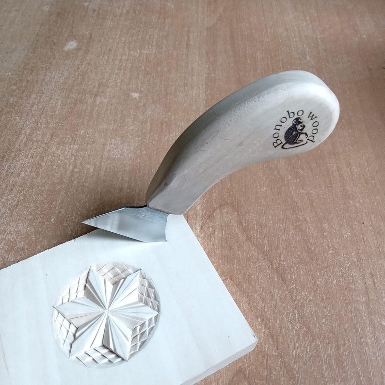 May include: A wood carving knife with a light-colored wooden handle and a silver blade is carving a star design into a piece of light-colored wood. The handle has the text "bonobo wood" and a monkey logo.