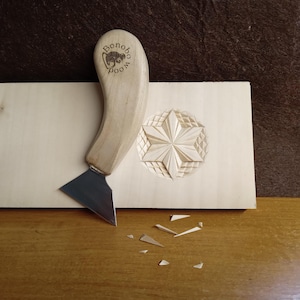 May include: A wood carving tool with a curved wooden handle and a silver blade rests on a light-colored wooden surface. A carved star design is visible on the wood, along with the text "Bonobo Wood". Small wood shavings are scattered nearby.