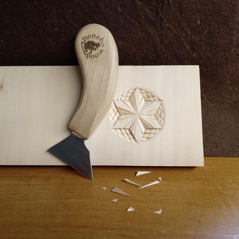 May include: A wood carving tool with a curved wooden handle and a silver blade rests on a light-colored wooden surface. A carved star design is visible on the wood, along with the text "Bonobo Wood". Small wood shavings are scattered nearby.