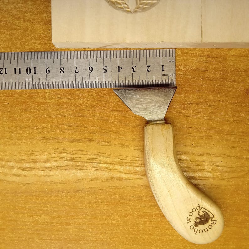 May include: A wood carving tool with a curved, light-colored wooden handle and a silver blade. The handle has the word "wood" and the word "Bonobo" printed on it. A ruler shows measurements in millimeters.