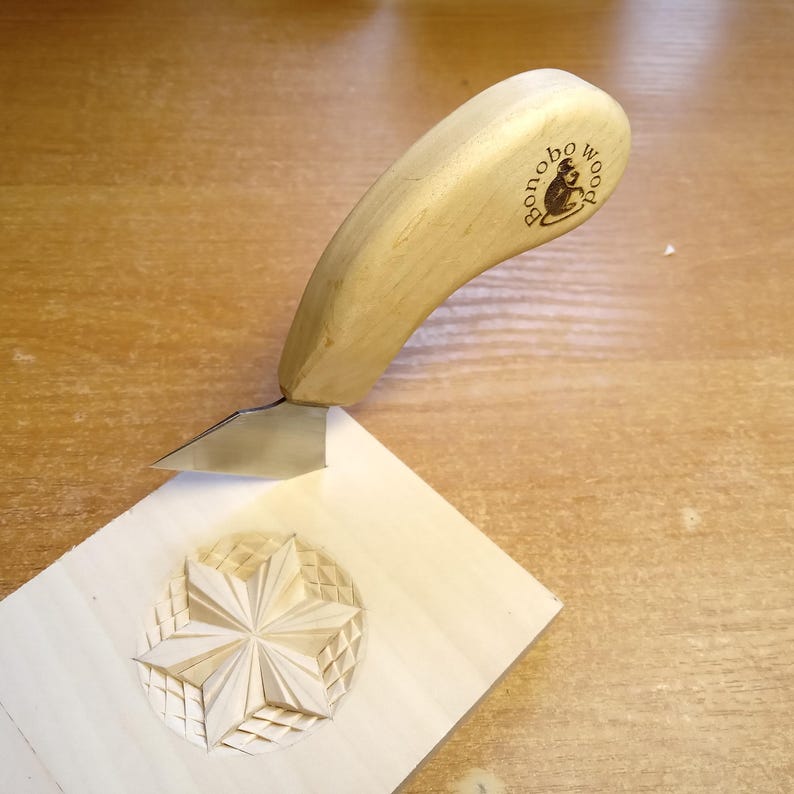 May include: A wood carving knife with a light-colored wooden handle is positioned above a carved wooden block. The block features a detailed star design within a circular border. The knife's blade is silver, and the handle has the text "bonobo wood" with a monkey logo.