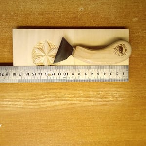May include: A wood carving knife with a curved wooden handle and a dark metal blade rests on a wooden block with a carved star design. A ruler shows measurements in inches. The handle is stamped with the text "Bonobo Wood".