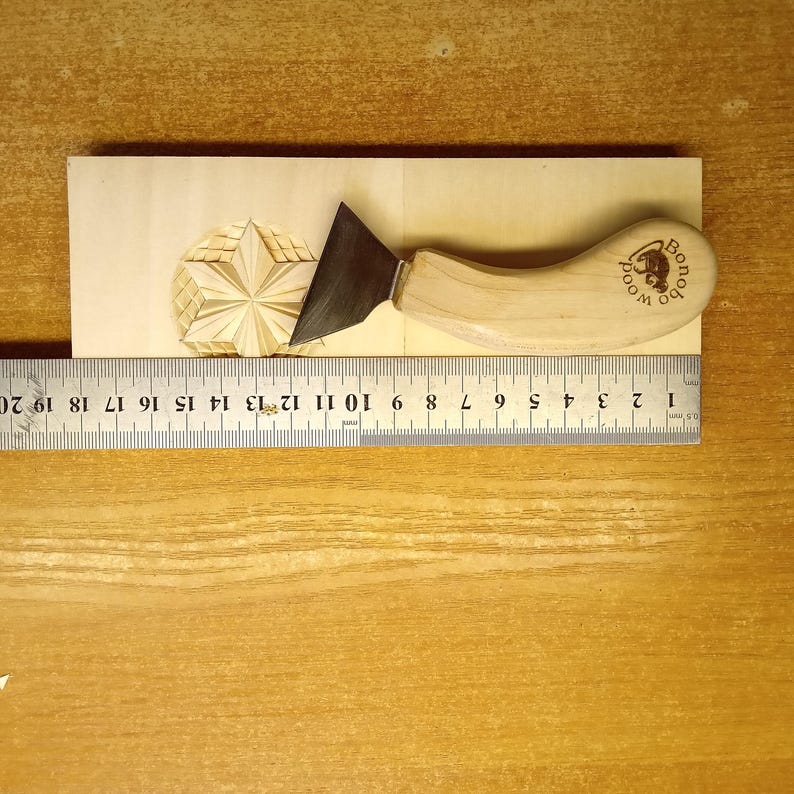 May include: A wood carving knife with a curved wooden handle and a dark metal blade rests on a wooden block with a carved star design. A ruler shows measurements in inches. The handle is stamped with the text "Bonobo Wood".