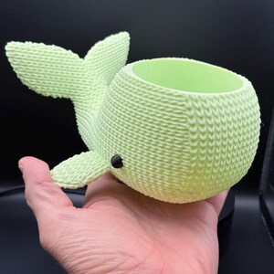 Puede incluir: Una maceta en forma de ballena de color verde claro con una apariencia texturizada y tejida. La ballena tiene un cuerpo redondo, una cola y aletas. Un pequeño punto negro representa el ojo. El interior es de un tono de verde más claro.