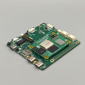 Könnte beinhalten: Eine grüne Raspberry Pi Compute Module 5 Leiterplatte mit verschiedenen elektronischen Komponenten, darunter Chips, Anschlüsse und Ports. Auf der Platine steht "Raspberry Pi Compute Module 5". Die Platine liegt auf einer grauen Oberfläche.