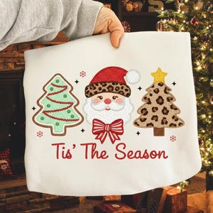 Può includere: Un cuscino bianco a tema natalizio con un design festivo. Presenta un Babbo Natale con un cappello leopardato, un albero di Natale verde e un albero di Natale leopardato. Il testo "Tis' The Season" è stampato sotto.