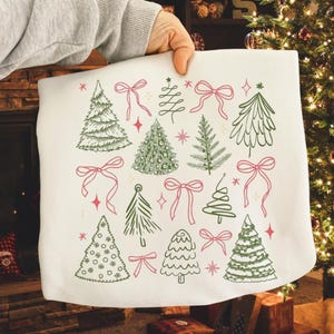 Può includere: Tessuto bianco con un design a tema natalizio. Il design presenta vari alberi di Natale verdi, fiocchi rosa e stelle rosse. Il tessuto è tenuto in mano da una persona.