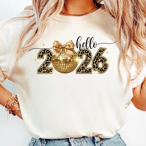 Può includere: T-shirt color crema con una palla da discoteca dorata, un fiocco dorato e i numeri "2026" in un motivo leopardato. La parola "hello" è scritta sopra i numeri in corsivo. Stelle dorate sono sparse intorno al design.