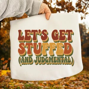 Può includere: Una borsa tote bianca con la scritta "LET'S GET STUFFED (AND JUDGMENTAL)" in stile retrò. Il testo è nei toni del marrone, arancione e giallo con contorni verdi. La borsa è tenuta contro uno sfondo autunnale sfocato.