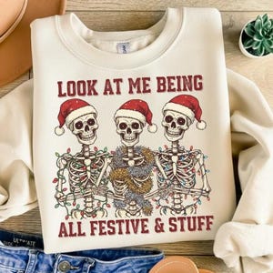 Può includere: Felpa color crema con una grafica di tre scheletri con cappelli di Babbo Natale e luci natalizie. Il testo "LOOK AT ME BEING ALL FESTIVE & STUFF" è stampato in rosso.