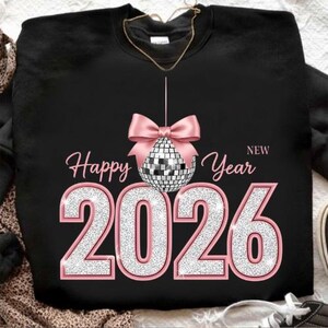 Può includere: Felpa nera con fiocco rosa e grafica a palla da discoteca. Le parole "Happy New Year 2026" sono visualizzate in rosa e glitter argento. Un design festivo per celebrare il nuovo anno.