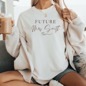 Può includere: Felpa color crema con la scritta "FUTURE Mrs. Smith" in corsivo nero. La felpa ha girocollo e maniche lunghe. È visibile una collana d'oro. La persona tiene in mano un bicchiere di caffè freddo.