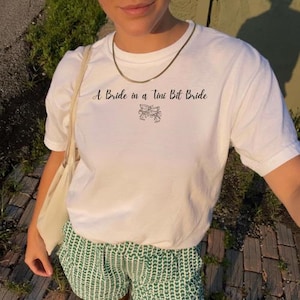 Può includere: T-shirt bianco con la scritta "A Bride in a Tiny Bit Bride" in corsivo nero. La maglietta presenta una piccola grafica a fiocco sotto il testo. La persona indossa pantaloncini verdi e bianchi e una collana d'oro.