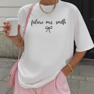 Può includere: T-shirt bianco con la scritta "future mrs. smith" in corsivo nero e un fiocco. La maglietta è abbinata a pantaloni a righe rosa e bianche e a una borsa rosa. Sono visibili anche una collana e braccialetti dorati.