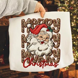Può includere: Borsa tote natalizia bianca con un Babbo Natale che ammicca, con cappello rosso e barba bianca. Le parole "Merry Christmas" sono ricamate con stampa leopardata e scritta rossa.