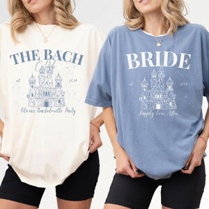 Può includere: Due magliette, una color crema e una blu, con testo grafico e illustrazioni di castelli. La maglietta color crema recita "The Bach" e "Olivia's Bachelorette Party". La maglietta blu dice "Bride" e "Happily Ever After". Entrambe le magliette hanno l'anno 2024.
