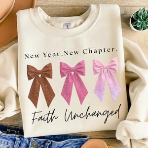 Può includere: Felpa color crema con la scritta "New Year. New Chapter. Faith Unchanged." Presenta tre fiocchi glitterati marroni, rosa e rosa sfumato. Un design casual e d'ispirazione.