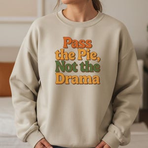 Può includere: Felpa beige con la scritta "Pass the Pie, Not the Drama" in arancione, verde e giallo. La felpa ha un girocollo e maniche lunghe.