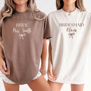 Può includere: Due magliette, una marrone e una bianca, con testo. La maglietta marrone dice "BRIDE Mrs. Smith" con una grafica a fiocco. La maglietta bianca dice "BRIDESMAID Olivia" con una grafica a fiocco. Entrambe le magliette sono a maniche corte.