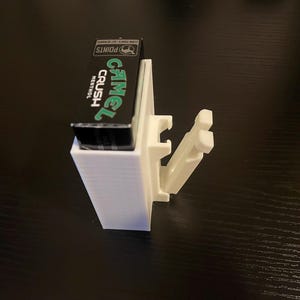 Puede incluir: Un soporte blanco para cigarrillos impreso en 3D con un paquete de cigarrillos Camel Crush Menthol negro y verde. El soporte tiene una base rectangular y un brazo articulado.