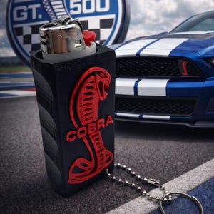 May include: Black lighter case featuring a red Cobra logo and text, attached to a keychain. A blue and white sports car is in the background, along with a GT 500 logo.