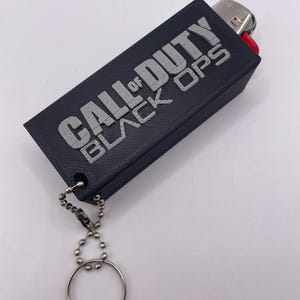 May include: Black rectangular lighter case with the text "CALL OF DUTY BLACK OPS" in silver lettering. The case has a silver key ring attached with a ball chain. The lighter is visible at the top.