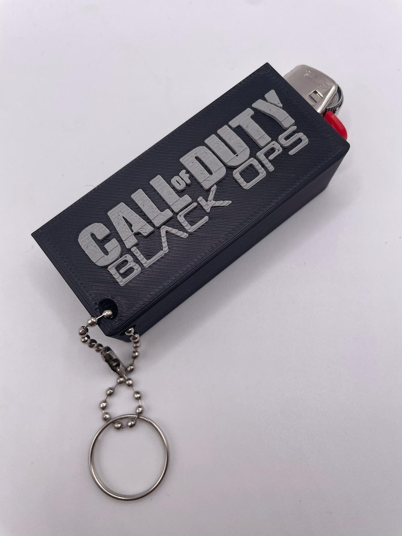 May include: Black rectangular lighter case with the text "CALL OF DUTY BLACK OPS" in silver lettering. The case has a silver key ring attached with a ball chain. The lighter is visible at the top.