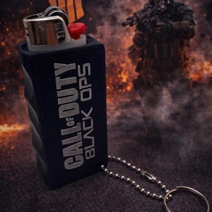 May include: A black lighter with a red ignition button is encased in a navy blue sleeve. The sleeve has the words "CALL OF DUTY BLACK OPS" printed on it in silver. A silver chain and keyring are attached to the sleeve.