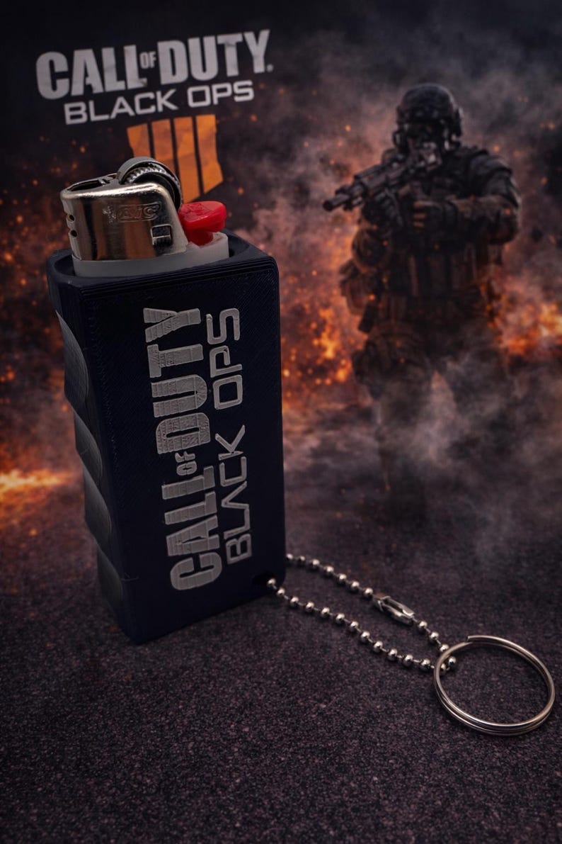 May include: A black lighter with a red ignition button is encased in a navy blue sleeve. The sleeve has the words "CALL OF DUTY BLACK OPS" printed on it in silver. A silver chain and keyring are attached to the sleeve.