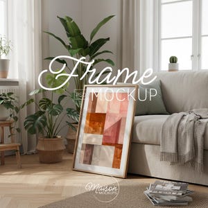 Könnte beinhalten: Gerahmter Kunstdruck-Mockup mit einem abstrakten geometrischen Design in Orange-, Rosa- und Brauntönen. Der Rahmen lehnt an einem beigen Sofa in einem hellen Raum mit Pflanzen. Der Text "Frame Mockup" ist sichtbar.