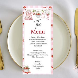Op de afbeelding: Een rechthoekige menukaart met een roze en wit geblokte rand, voorzien van aardbei-illustraties, een waslijn-afbeelding met babyartikelen en de tekst The Menu, gevolgd door lijsten met eten en drinken, waaronder kipsalade en thee.