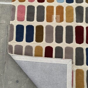 Puede incluir: Una alfombra rectangular con un patrón repetitivo de rectángulos verticales coloridos. Los rectángulos son en tonos de rosa, azul, marrón, gris y dorado, sobre un fondo crema. Se ve la parte inferior de la alfombra, mostrando una tela gris.
