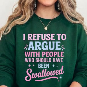 K&ouml;nnte beinhalten: Dunkelgr&uuml;nes Sweatshirt mit dem Text "I REFUSE TO ARGUE WITH PEOPLE WHO SHOULD HAVE BEEN SWALLOWED" in Rosa und Hellblau. Das Sweatshirt hat einen Rundhalsausschnitt und lange &Auml;rmel.