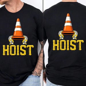May include: Black t-shirt featuring a graphic of an orange and white traffic cone held by yellow and white gloved hands. The word "HOIST" is printed in yellow, stylized lettering below the graphic.