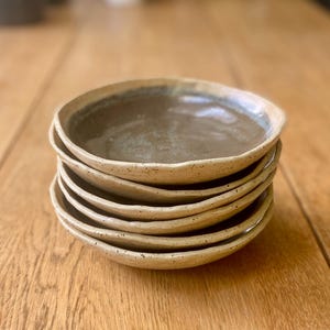 May include: A stack of six handmade ceramic plates. The plates have a speckled, beige exterior and a darker, brownish-grey interior. The rims have a slightly irregular, wavy edge. The plates are stacked on a wooden surface.